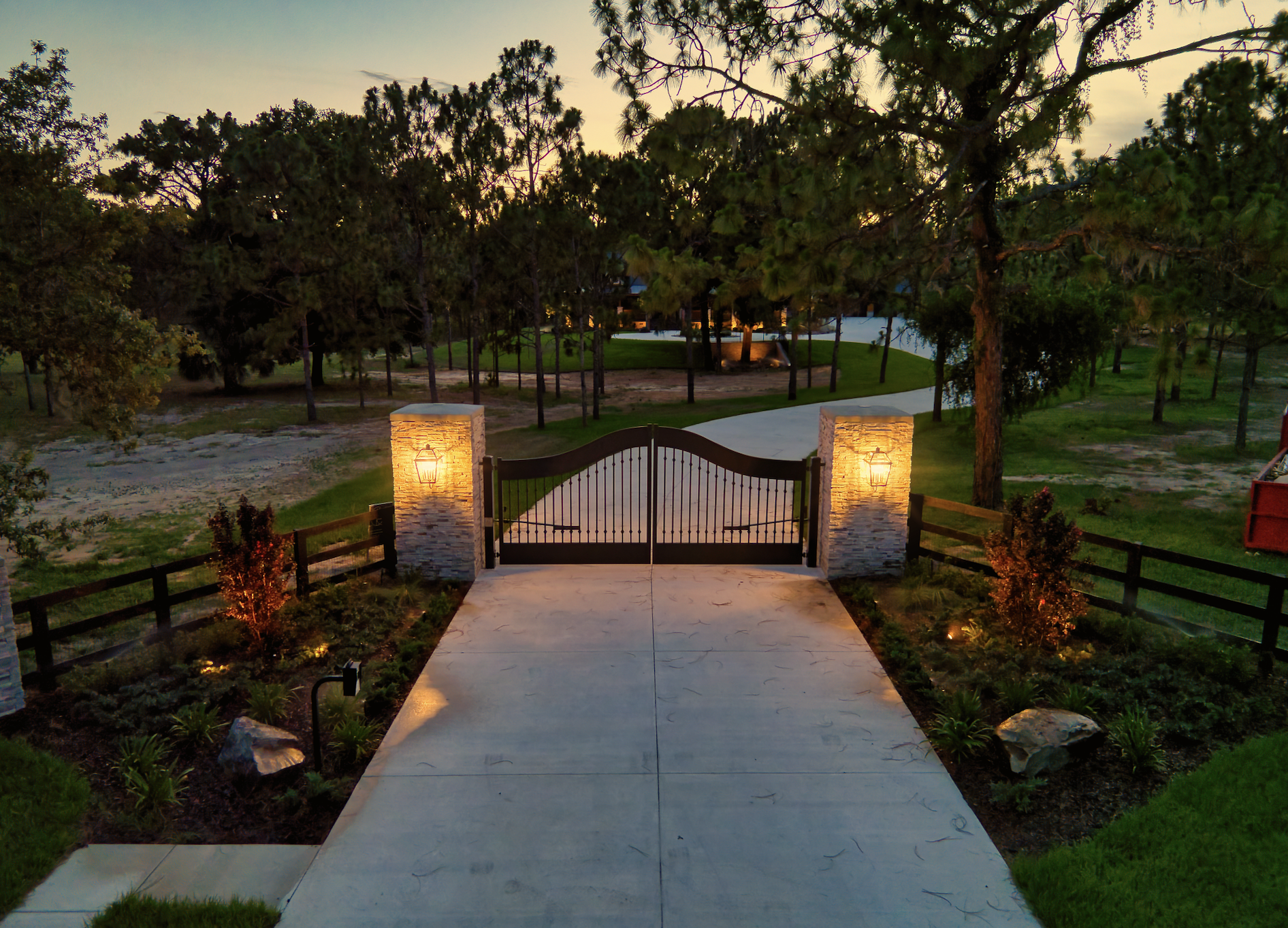 Illuminated gated driveway at dusk with trees.