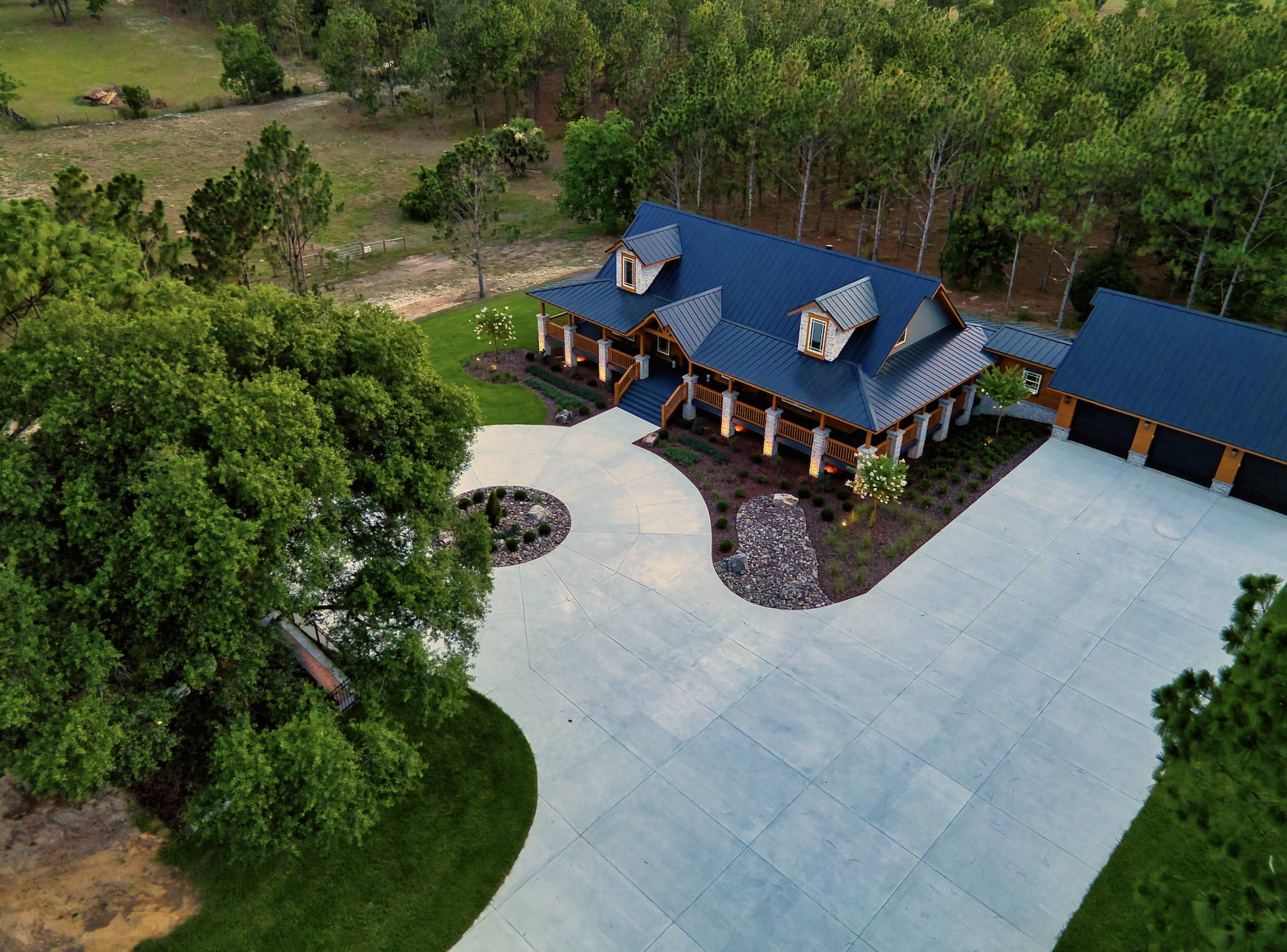 Aerial view of a custom home with a curved concrete driveway, metal roof, and surrounding trees.