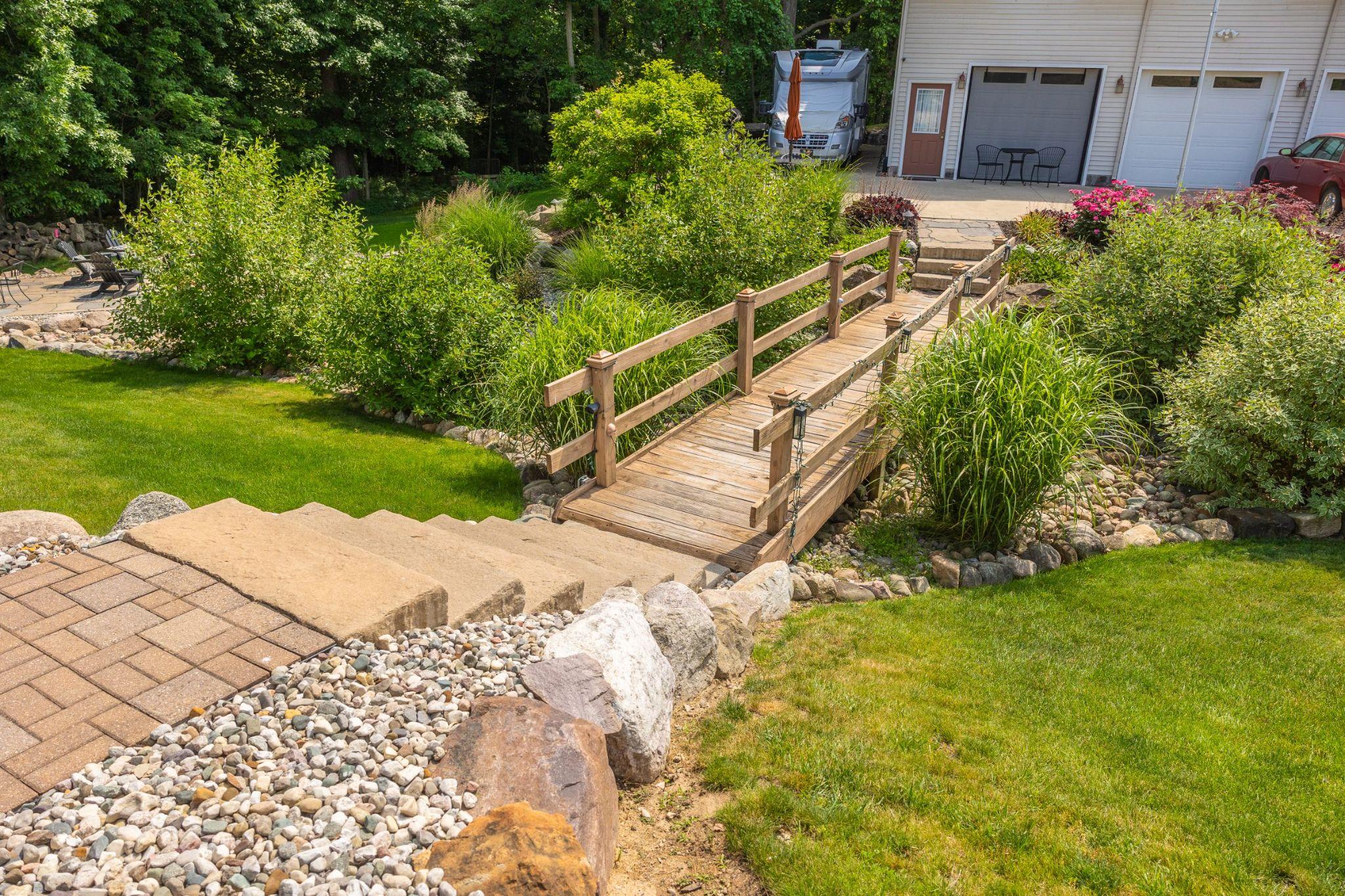 Backyard garden featuring a wooden footbridge over decorative stone, surrounded by lush planting and a paved walkway.