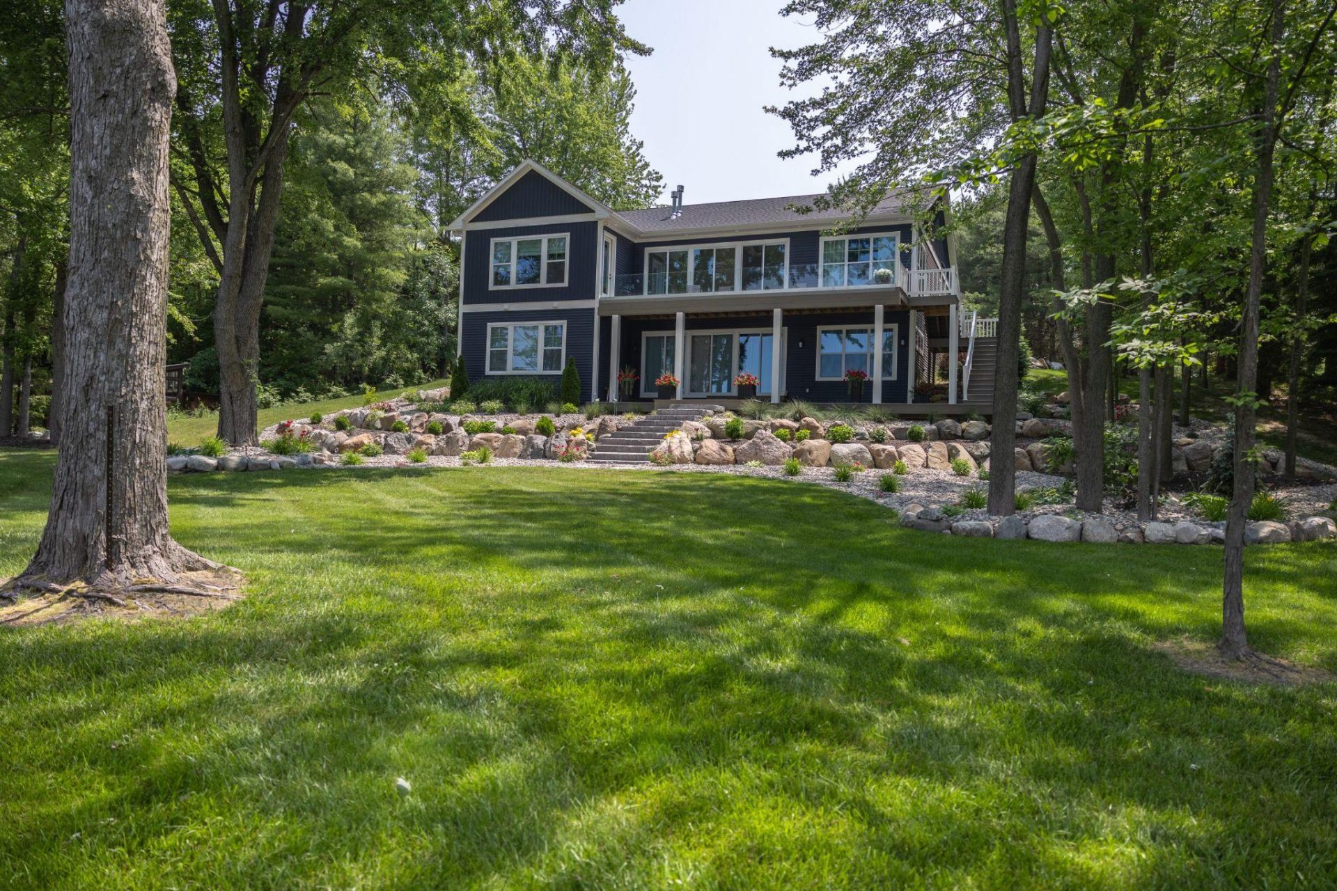 Modern home set on a sloped landscape with stone retaining walls, wide lawn, and mature trees providing shade.