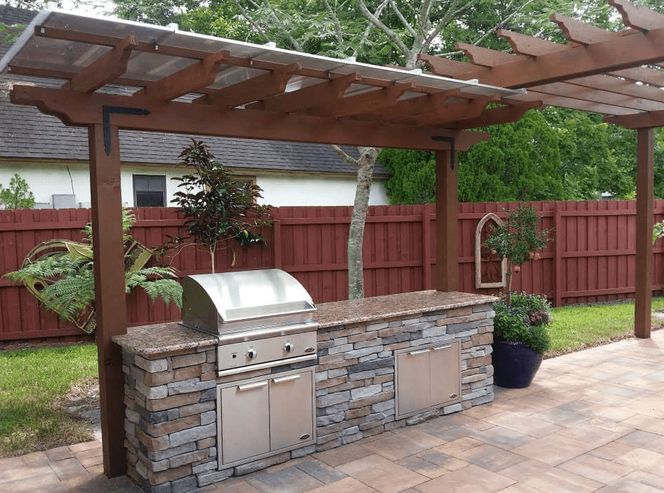 Outdoor kitchen with built-in grill, stone cabinetry, and wooden pergola in a fenced backyard.