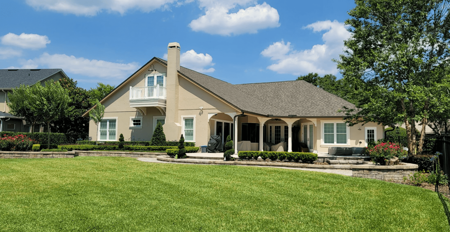 Rear exterior of a home with expansive green lawn, patio seating, and mature trees.