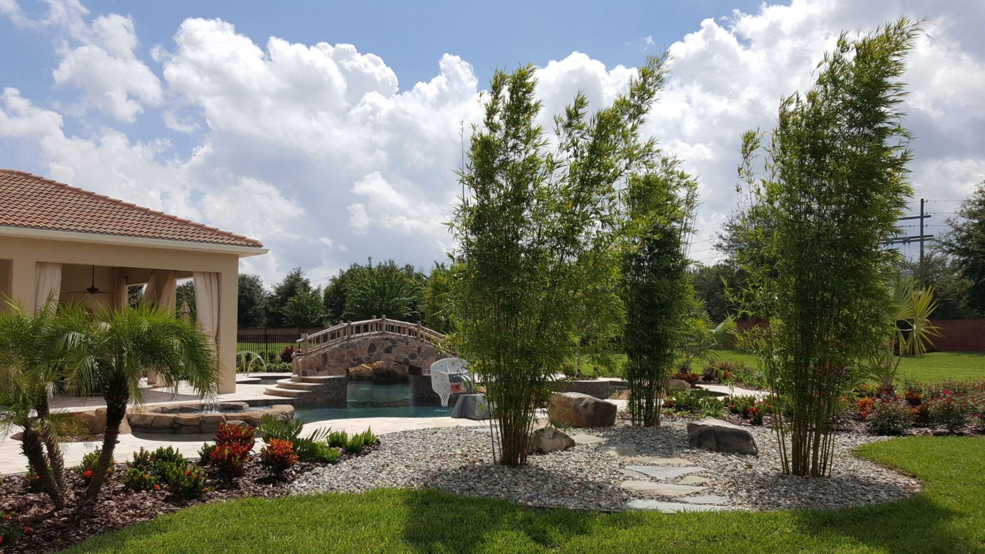 Resort-style backyard with pool, stone bridge, bamboo plantings, and decorative rock landscaping.