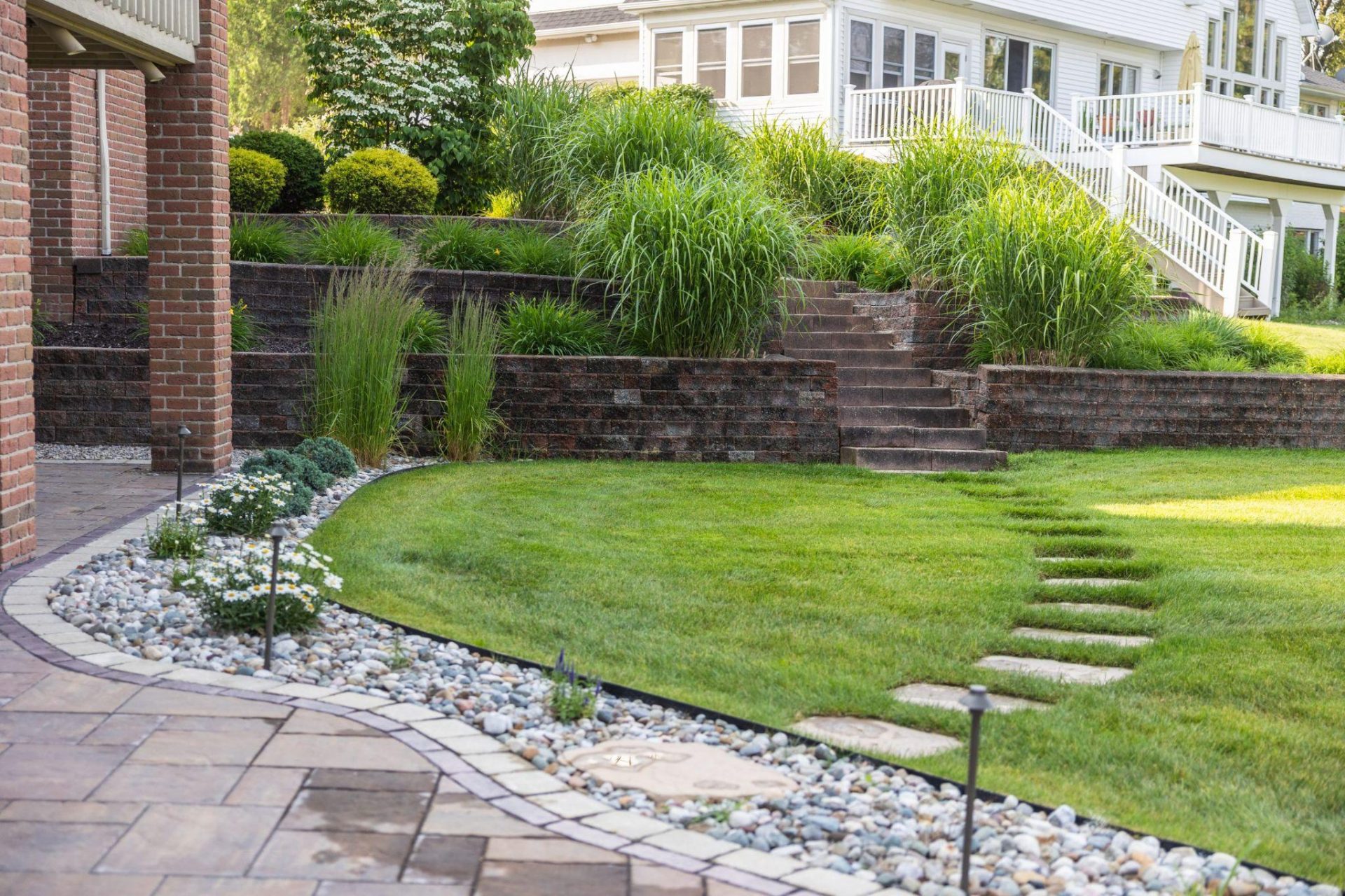 Tiered retaining walls with stone steps, ornamental grasses, and a curved lawn creating a structured backyard landscape.