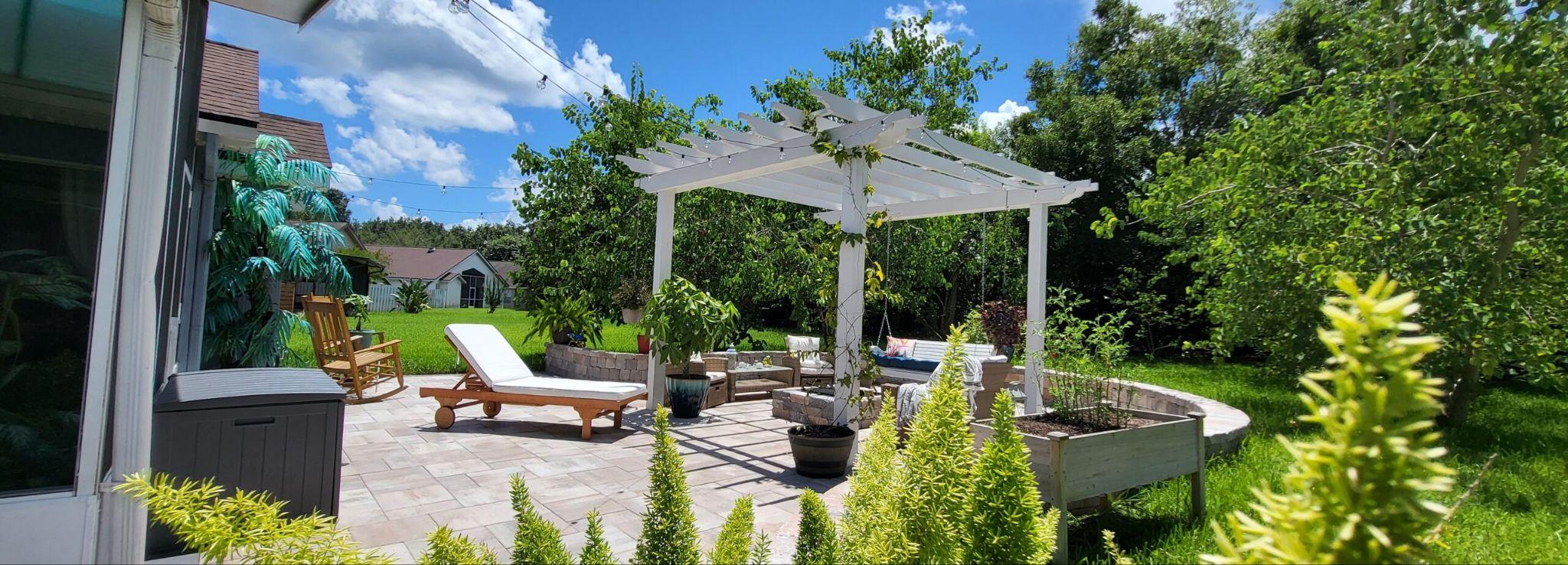 White pergola over a paved patio with lounge chairs, raised planters, and surrounding greenery.